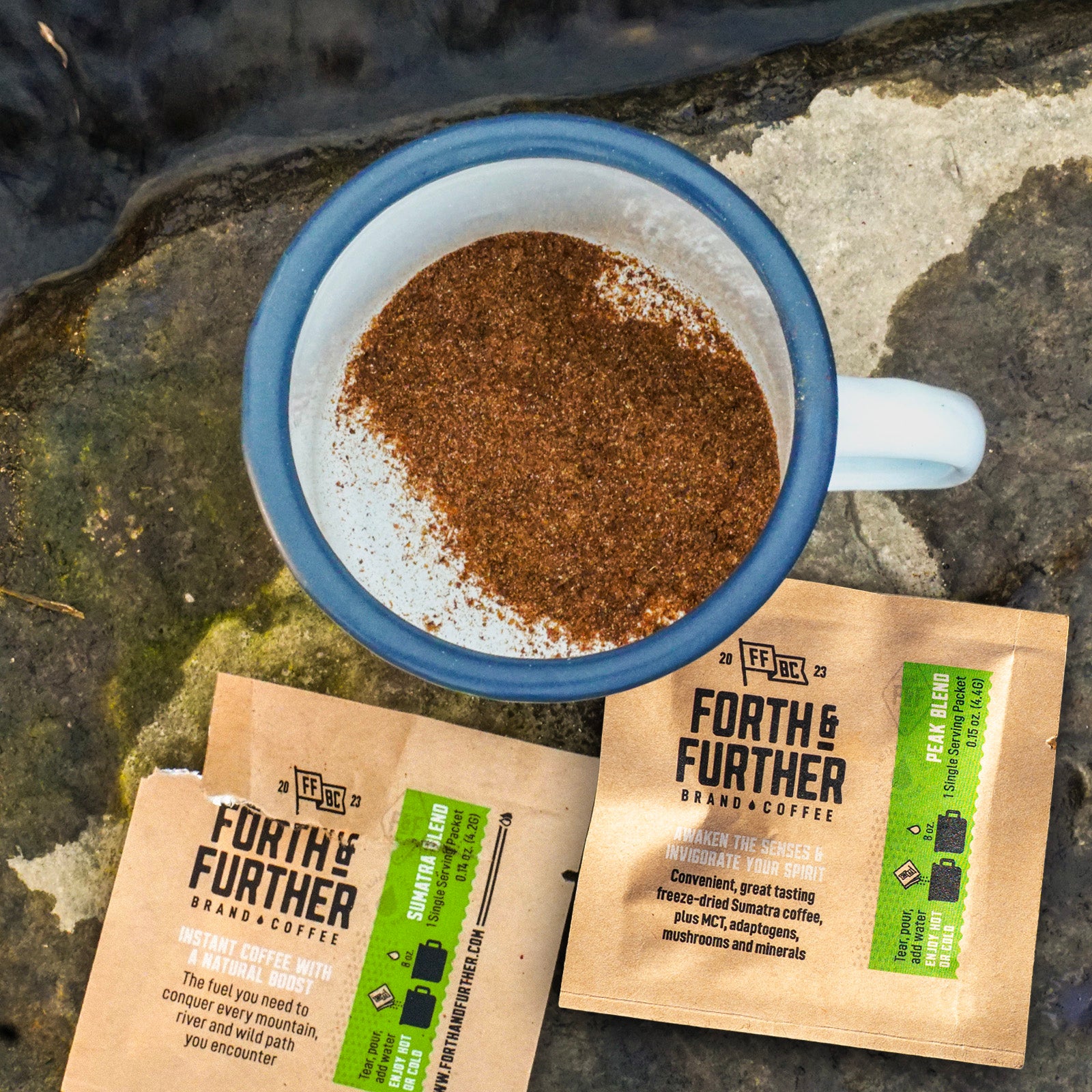 Camp mug with freeze-dried coffee next to Forth & Further coffee packets on a stone next to a creek