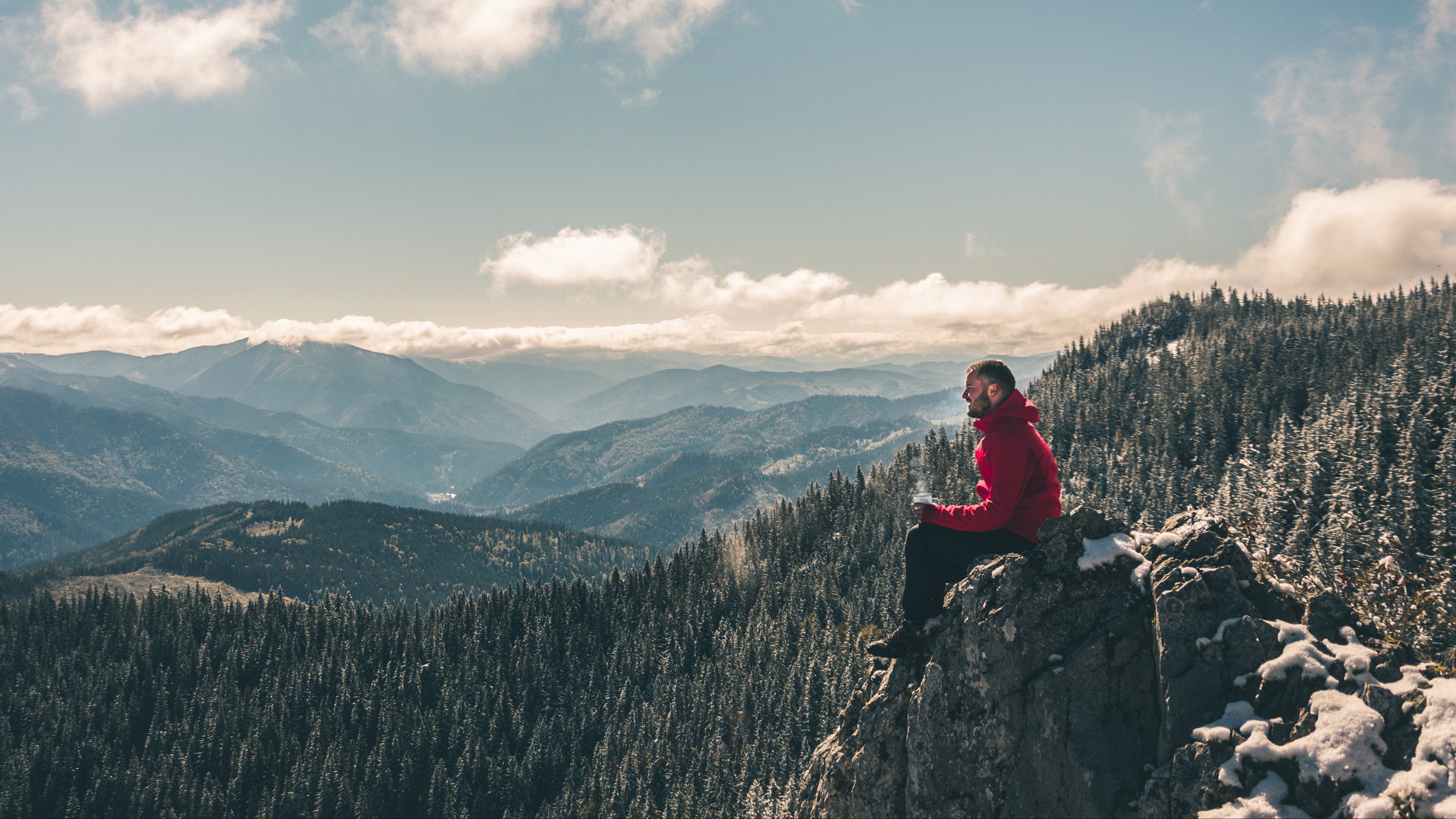 Person in a red jacket holding a mug of hot coffee sitting on a mountain top with a scenic view of trees and mountains